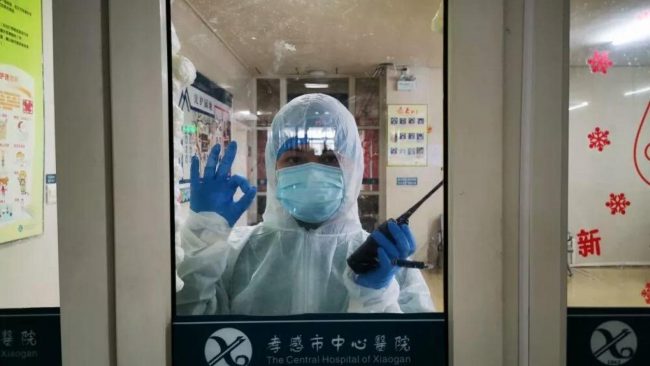 Healthcare worker in personal protective equipment gives an okay hand signal from behind a hospital door